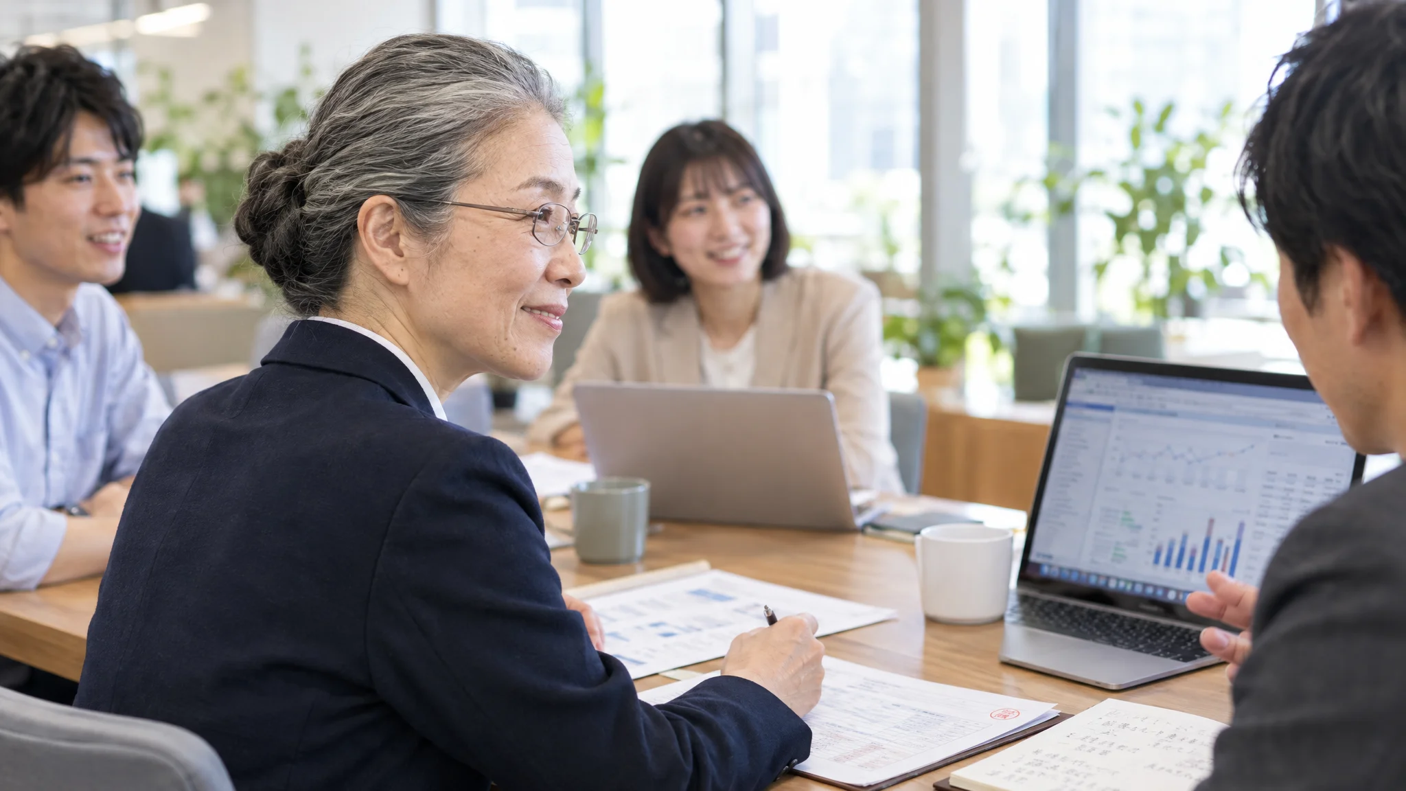 In a quiet office space, a Japanese executive sits at a desk, intently watching an organizational analysis dashboard displayed on a monitor. The screen shows engagement scores for each department as a color-coded heatmap, with a yellow caution mark on a specific area. Potted plants are placed around, depicting a calm business environment.