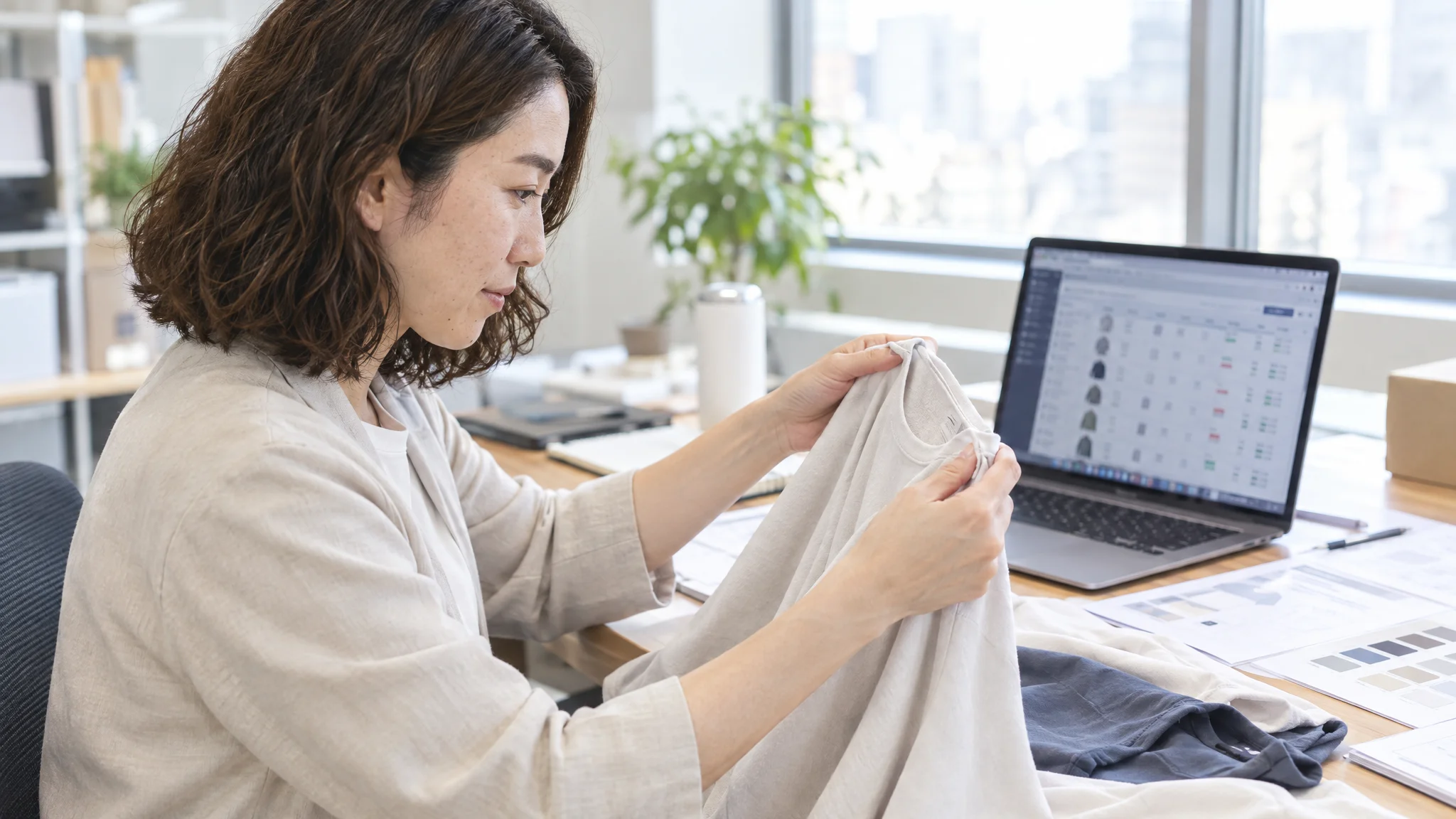 日本のIT企業のオフィスで、日本人のデータアナリストが真剣な表情でPC画面に向かっている。画面には、AIが検品時に抽出した商品の色相分布（ヒストグラム）と、マスターデータとの偏差を示す散布図が詳細に表示されている。デスクの上には、解析対象となるアパレル生地のサンプルと、日本語で書かれた技術仕様書が置かれている。室内の光は一定に保たれ、プロフェッショナルな分析環境が描写されている。