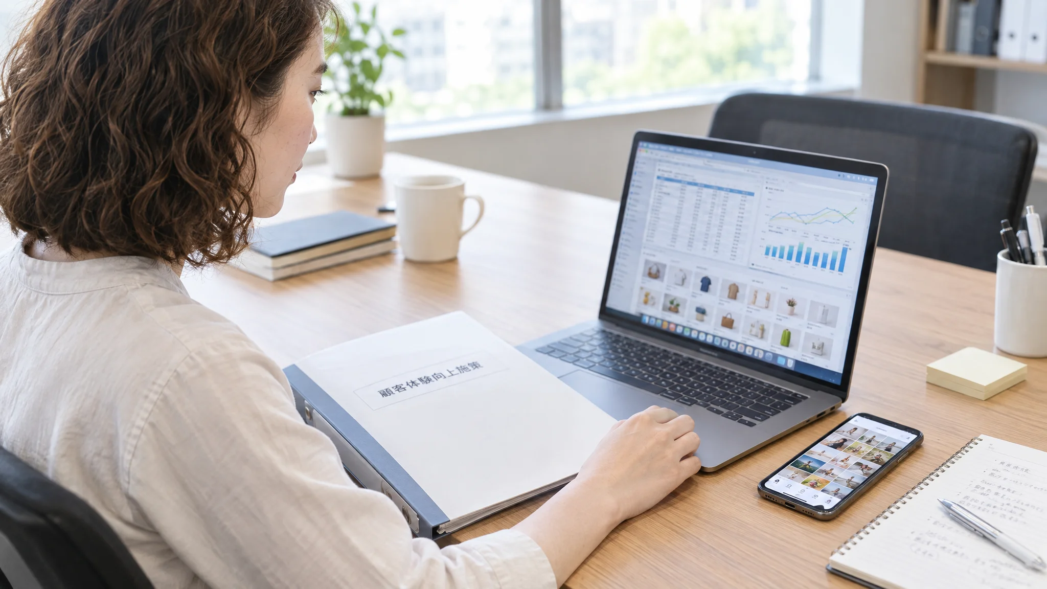 A bright Japanese office environment. On the desk, there is a binder labeled "Customer Experience Improvement Measures" in Japanese and an actual smartphone. The smartphone screen shows the landing page of an in-house EC site, beautifully displaying customer Instagram posts in a carousel format. No people are shown, conveying the atmosphere of an organized business setting.